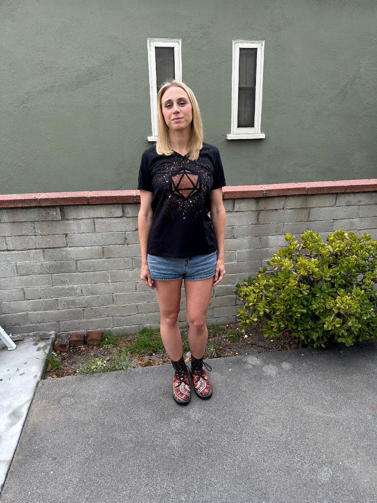 Amy Vorpahl wearing jean shorts, patterned combat boots, and a black tshirt with a D20 die design made out of bleach splatters