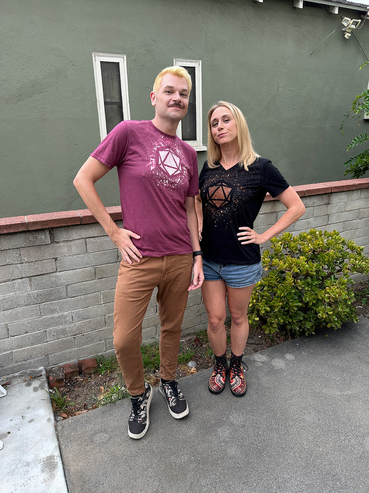 Amy Vorpahl standing with her hand on her hip. She's wearing a black tshirt with a D20 die design made out of bleach splatters. Xander Jeanneret is standing next to her wearing a coral colored shirt with the same D20 bleach design.