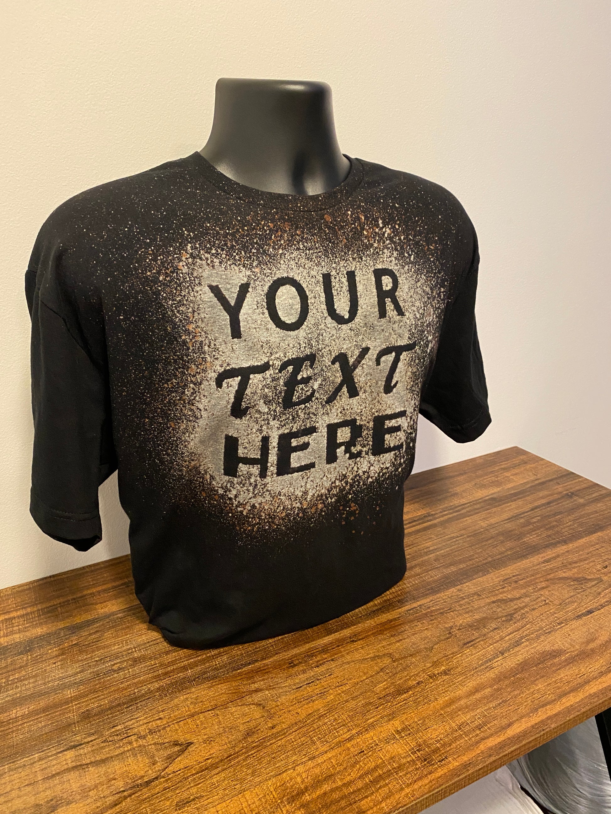 Black shirt that says "your text here" on it, made with bleach. The shirt is on a shirt form, which is sitting on a wood table.