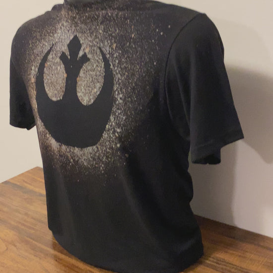 A black shirt on a shirt form with a bleach design that shows the Rebel Alliance logo from Star Wars. There is a pixel heart on the back of the shirt, which is also made with bleach splatter.