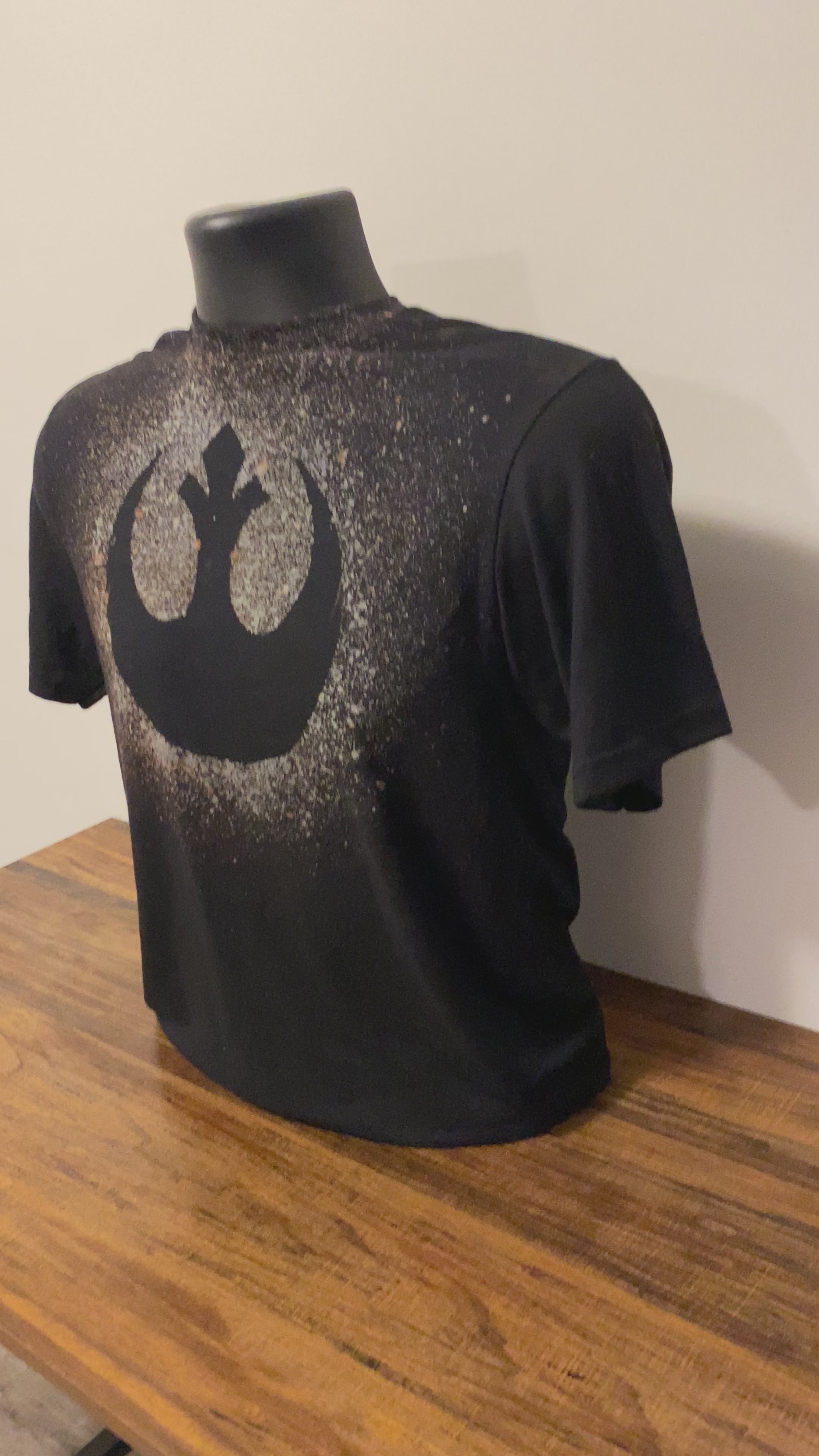 A black shirt on a shirt form with a bleach design that shows the Rebel Alliance logo from Star Wars. There is a pixel heart on the back of the shirt, which is also made with bleach splatter.